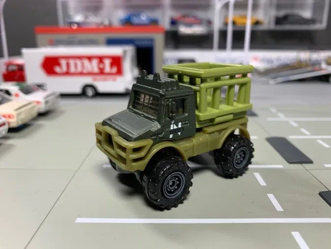 Un jouet Matchbox vert représentant un Mercedes-Benz Unimog U1300 de la série Jurassic World est posé sur une surface grise avec d'autres véhicules en arrière-plan.