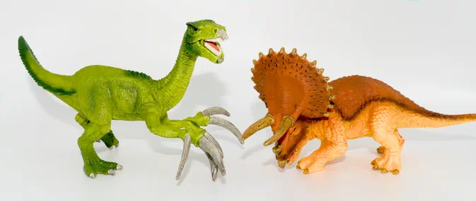Deux figurines de dinosaures en plastique : un théropode vert aux griffes levées face à un tricératops orange, sur fond blanc.