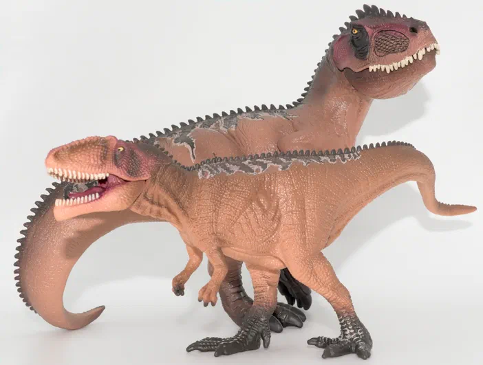 Deux figurines de dinosaures bruns avec des crêtes grises et des dents blanches posées sur un fond blanc.