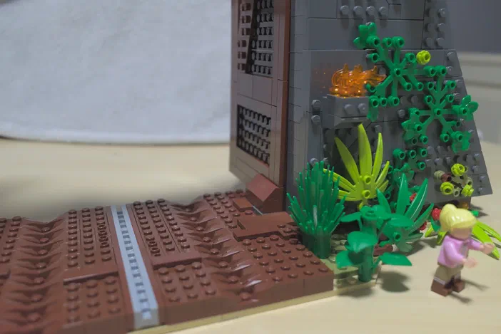 Un ensemble de construction LEGO Jurassic World montrant un minifigurine blonde près d'une structure grise avec des plantes vertes et une base brune.