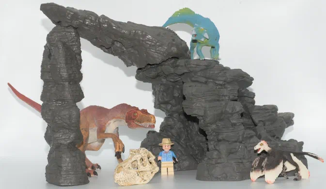 Un ensemble de jouets incluant des dinosaures, un squelette et une figurine Lego devant une structure rocheuse.