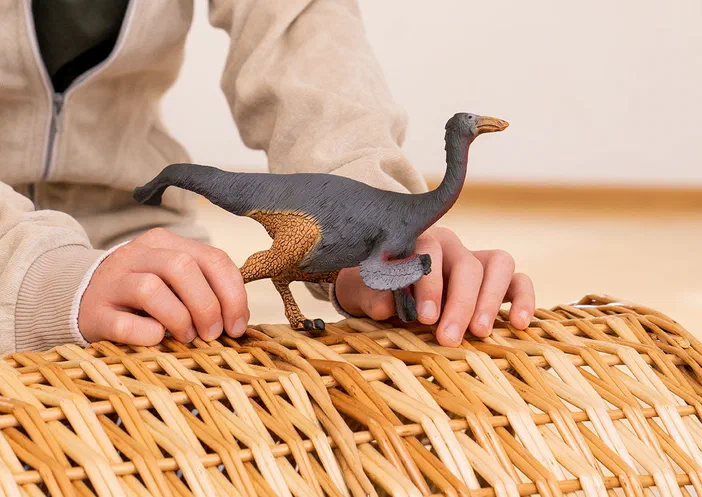 Une main tient un jouet de dinosaure Gallimimus sur une surface en vannerie.