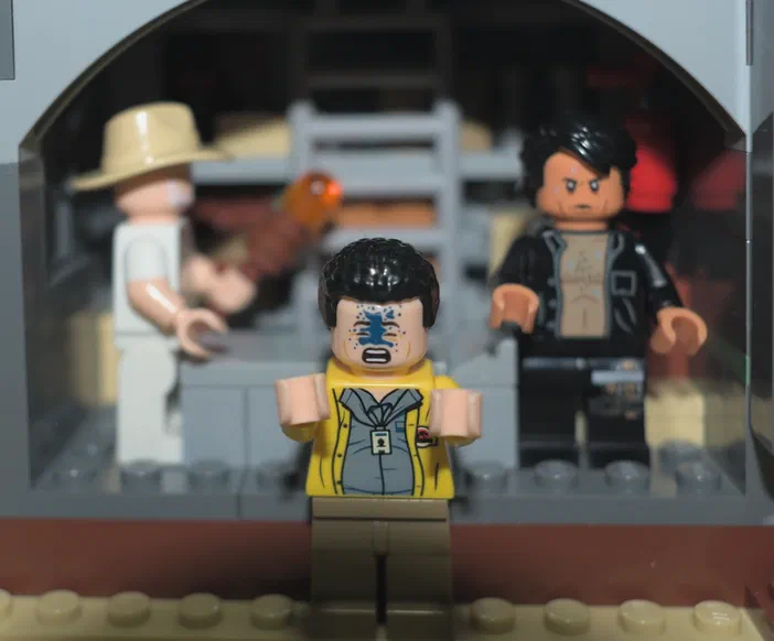 Trois figurines LEGO dans un décor de Jurassic World : une avec des bleus sur le visage au premier plan, et deux autres en arrière-plan.