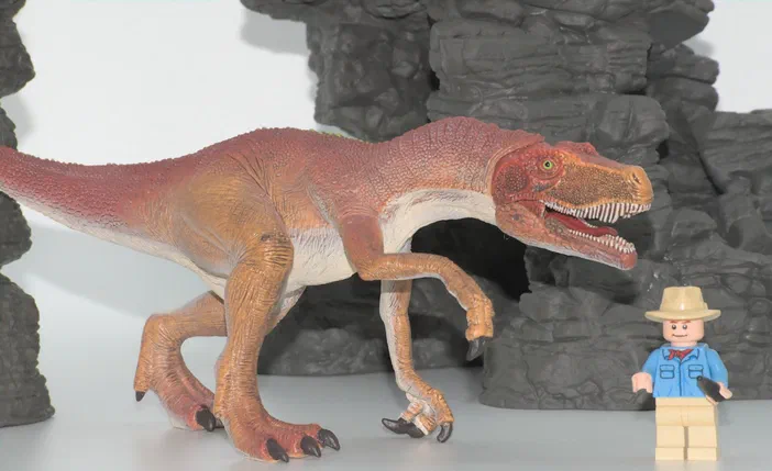 Un jouet de dinosaure rouge et beige se tient devant un minifigurine LEGO habillée d'un chapeau, dans une scène avec des rochers gris.